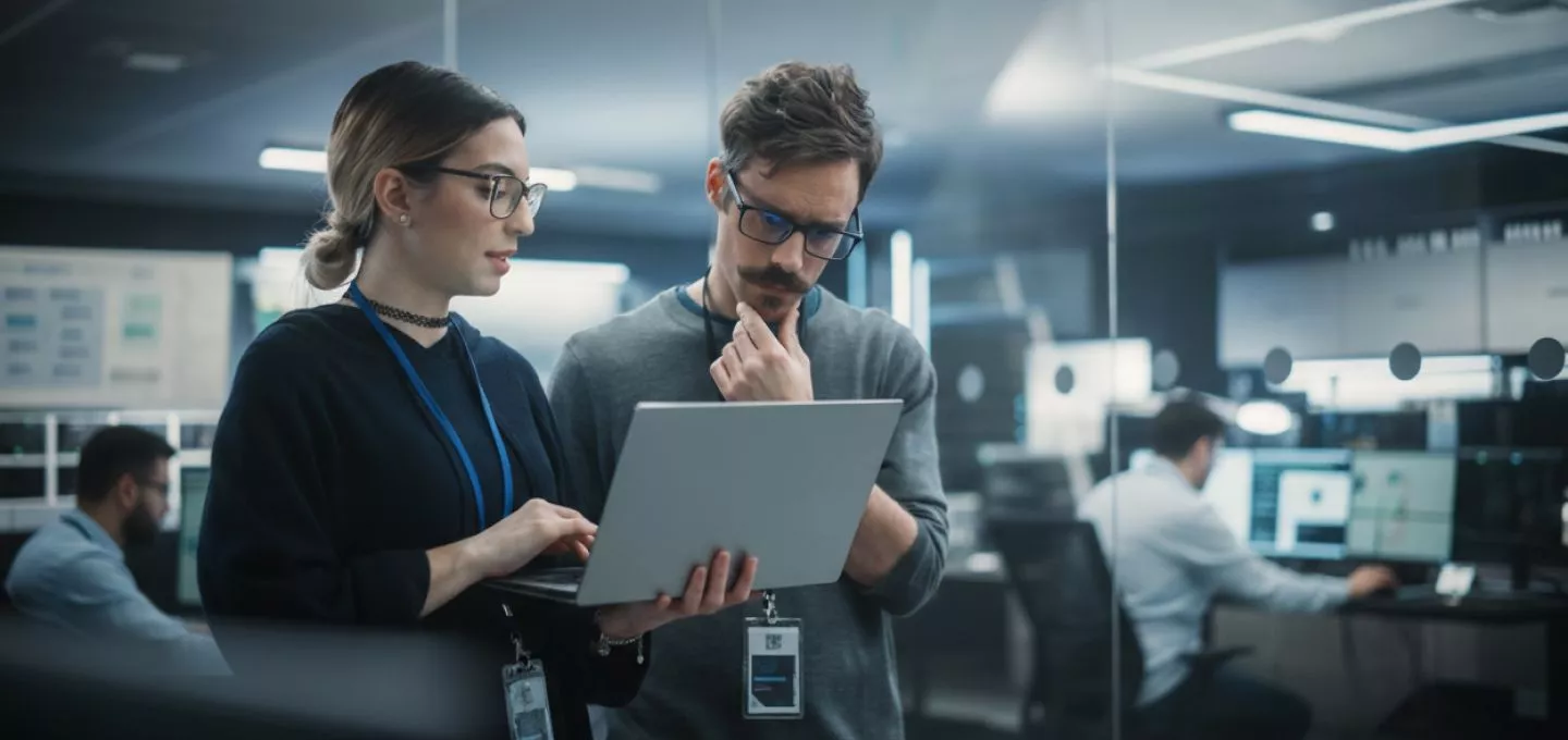  A photograph set in a modern, busy IT command center or data office. Two professionals, a woman and a man, are standing and closely reviewing information on a silver laptop the woman is holding. The woman wears glasses and an ID badge, pointing at the screen. The man, also wearing glasses and a mustache, looks intently at the screen with his hand on his chin, suggesting contemplation. In the blurred background, other colleagues are visible working at computer stations, emphasizing a collaborative and high-tech work environment, possibly for IT support or operations.