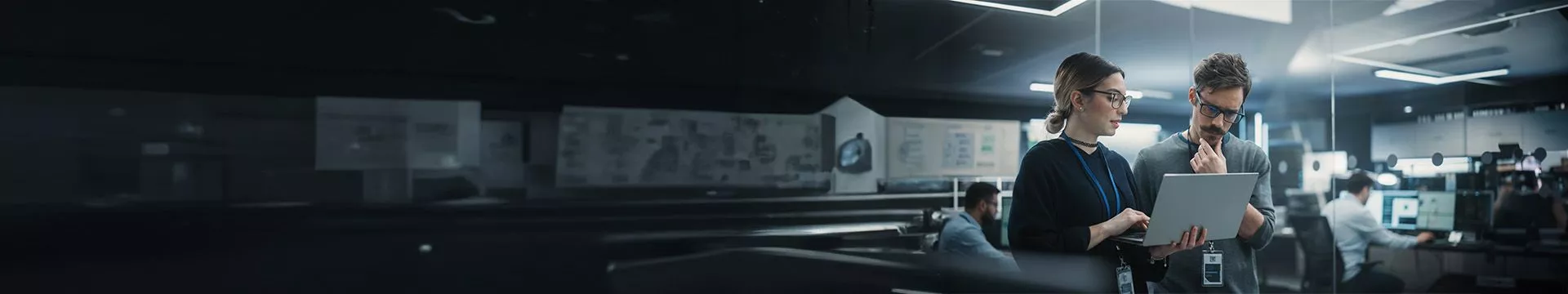 Dos profesionales de TI, una mujer con un portátil y un hombre, colaboran para resolver un problema en una oficina de soporte técnico. Simboliza el servicio de Gestión de TI o IT Total Care.