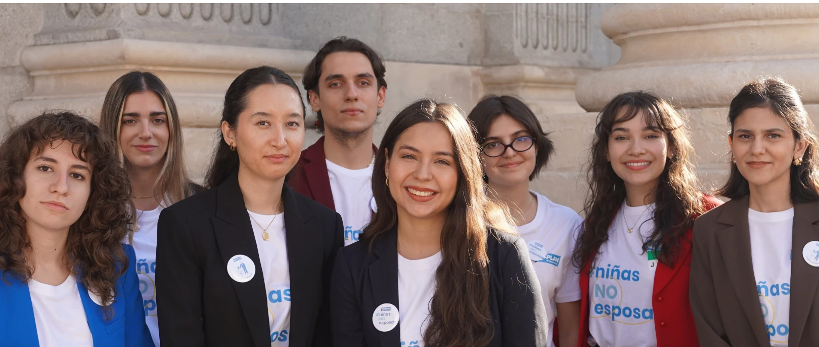 Photograph of a diverse group of eight young adults standing outdoors in front of a stone building, smiling and facing the camera. Several individuals wear buttons and shirts with text, including one that reads "niñas NO esposas," indicating a social or advocacy context.