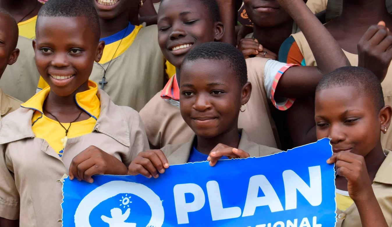 Photograph of a group of smiling children wearing beige uniforms holding blue signs with white "PLAN INTERNATIONAL" logos. The setting suggests a school or community event promoting Plan International's mission to support children's rights and equality.